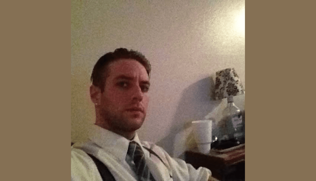 Man wearing a white shirt, striped tie, and suspenders in a dimly lit room.