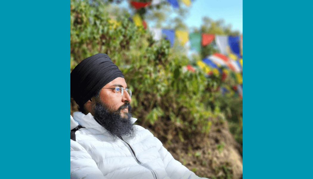 Bearded man wearing glasses, black turban, and white puffer jacket with colorful prayer flags behind.