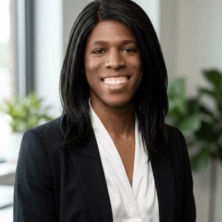 Smiling professional with long black hair wearing black blazer and white blouse in an office.
