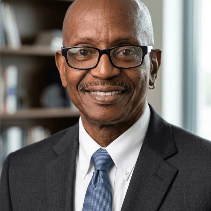 Smiling bald Black man with glasses, mustache, and hoop earring wearing a suit and tie.