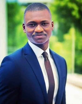 Smiling Black man in a blue suit and brown tie against blurred green foliage.