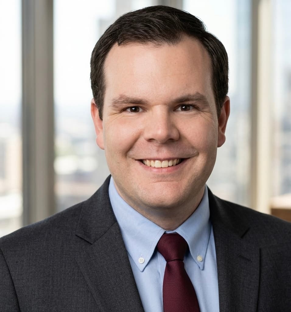 Smiling man with dark hair in a grey suit, light blue shirt, and maroon tie.
