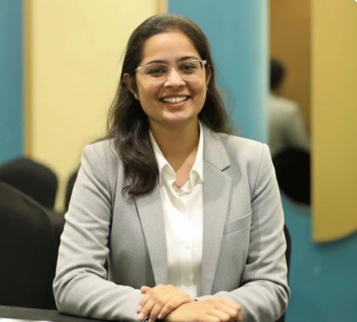 Smiling woman with glasses wearing a grey blazer and white shirt, sitting at a desk.