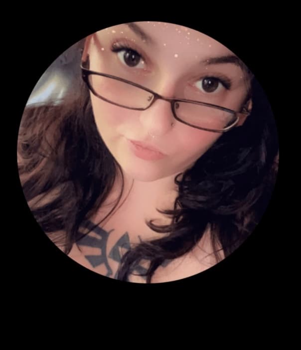 Woman with glasses, long dark hair, and a chest tattoo in a circular portrait.